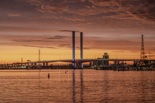 A dramatic sunset over Bolte Bridge in Melbourne, Australia. The sky transitions from deep blue to fiery orange, reflecting on the calm water.