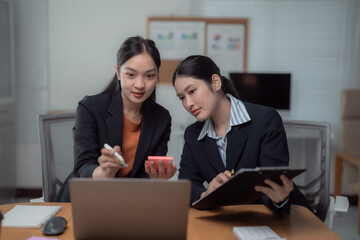 Two asian businesswomen are having a meeting in the office, using a laptop and taking notes on a clipboard, collaborating and discussing work together