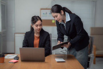 Two Asian businesswomen collaborating in a modern office, one working on a laptop while the other takes notes on a clipboard, engaged in a productive discussion and planning session