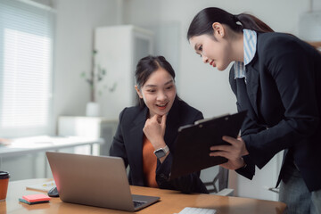 Two young businesswomen are discussing and working together using laptop and clipboard, analyzing financial statistics displayed on the laptop screen while sitting at desk in modern office