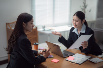 Two asian businesswomen are discussing about a document during a business meeting, one of them seems upset and is explaining something to her colleague who's taking notes