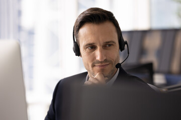 Positive thoughtful contact center manager man in wireless headset sitting at computer monitor in...