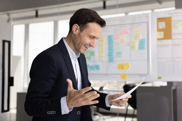 Cheerful excited Hispanic businessman making winner hand gesture, reading paper legal document with good news, financial report, smiling, laughing, celebrating professional career achievement