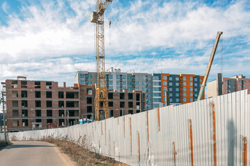 Construction site with a tall yellow crane, unfinished brick building, metal fence, and modern apartment buildings in the background under a blue sky with clouds