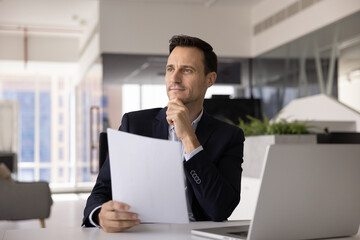 Positive thoughtful Latin legal professional man thinking on document at workplace, holding paper, touching chin, looking away. Business CEO, executive planning future deal