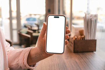 Close-up of a person holding a smartphone with blank white screen in a cozy cafe interior. Wooden counter and window background. Modern lifestyle, mockup for app or website