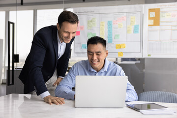 Happy Asian business professional man showing work result to boss standing nearby, sitting at laptop, typing, smiling. Multiethnic colleagues analyzing online product on computer