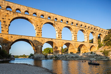 Fototapeta premium Pont du Gard, ancient Roman aqueduct bridge, Provence late afternoon golden light on arches mirroring in river, Gorges du Gardon, natural landscape designated Biosphere Reserve by UNESCO, France
