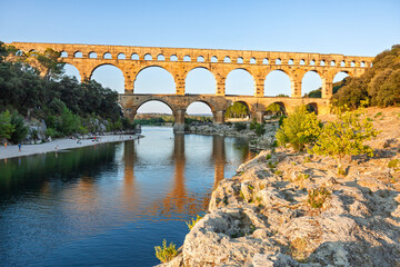 Fototapeta premium Pont du Gard, ancient Roman aqueduct bridge, Provence late afternoon golden light on arches mirroring in river, Gorges du Gardon, natural landscape designated Biosphere Reserve by UNESCO, France