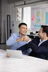 Two happy diverse business partners and friends enjoying effective productive teamwork, giving fist bump at table with tablet computer, celebrating group achievement, successful partnership