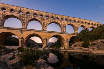 Pont du Gard, ancient Roman aqueduct bridge, colorful distant sunrise, under Provence starry night sky, Gorges du Gardon, natural landscape designated Biosphere Reserve by UNESCO, France