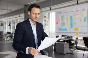 Focused Latin business executive man reading paper legal document, formal letter in office space alone, analyzing financial sales report, statistic data, doing paperwork, making decision