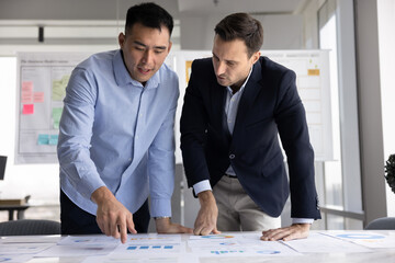 Two diverse male business colleagues brainstorming on project statistic data together, talking at table with paper marketing financial reports, discussing investment strategy