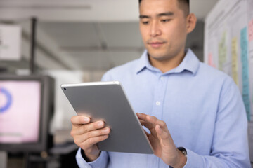 Focused young Asian business professional man using Internet technology on tablet for web communication, working on online project, financial statistics, analytic report