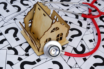 Stethoscope and damaged wooden house model on question marks background.