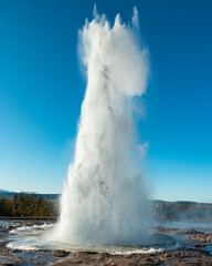 Strokkur geyser in Iceland erupts, shooting boiling water high into the air. A popular attraction in the Golden Circle, showcasing Iceland’s geothermal activity on a clear day with a blue sky. 