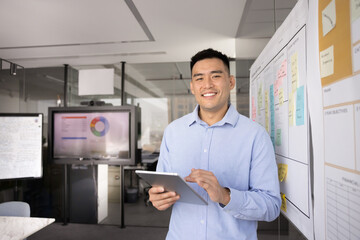Happy young Asian businessman holding digital tablet, standing in office workspace alone, looking...