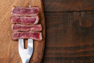 Carving fork with pieces of delicious beef on wooden table, top view. Space for text