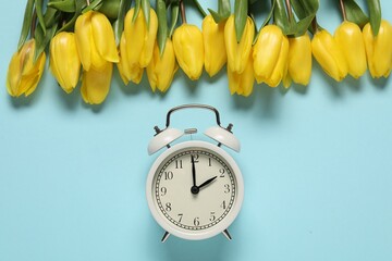 Spring time. Alarm clock and beautiful tulips on light blue background, flat lay