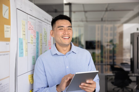 Positive successful Asian project manager working with tablet at white board with post it stickers, holding digital device, looking away, smiling, laughing, enjoying modern Internet technology for job