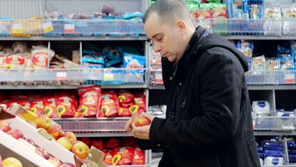 Man carefully picking ripe, red apples from grocery store produce display, examining freshness among colorful fruits and vegetables
