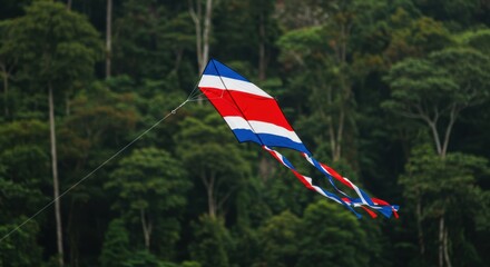 Fototapeta premium ?colorful kite with red, white, and blue stripes flying against lush forest backdrop?