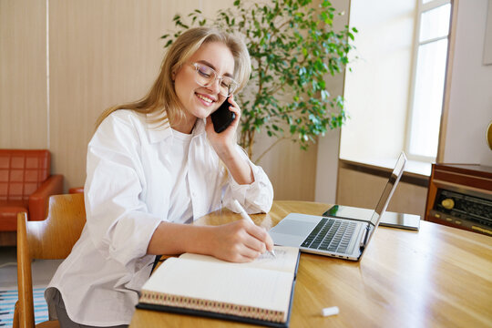 Young woman engaged in a phone call while taking notes at a wooden table in a well-lit room