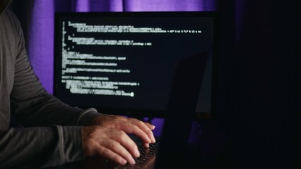 Freelancer quickly types computer code on keyboard which runs on freelancer's monitor in white font. Soft purple light in background. Freelancer working remotely from home late at night, side view