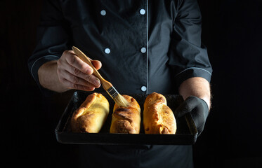 A skilled chef holding a tray of three golden rolls applies a glossy glaze with a brush. The...