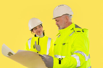 Construction workers examine blueprints in bright safety gear, discussing plans for a building project on yellow background