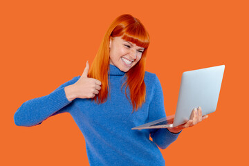 Happy woman giving thumbs up while using laptop against a bright orange background during a productive work session