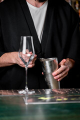 A bartender in a black robe prepares a cocktail using a shaker. The focus is on the skilled hands and the elegant glass ready for serving, showcasing the art of mixology.