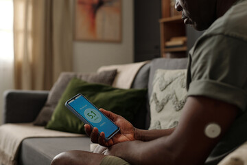 African American man checking blood glucose level using smartphone app while sitting in living room with cushion and bookshelf