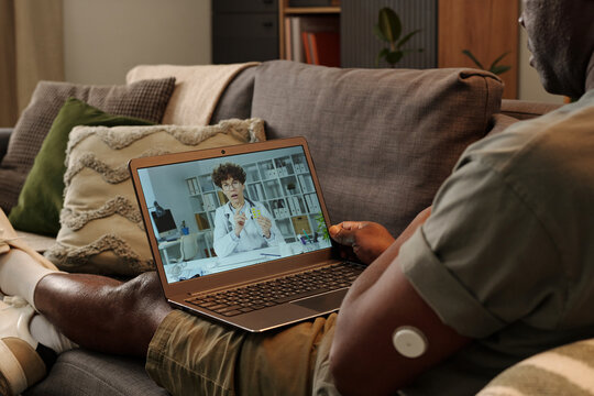 Person sitting on sofa in living room, using laptop for virtual medical consultation with doctor discussing health issues and treatment options, tech device visible on arm