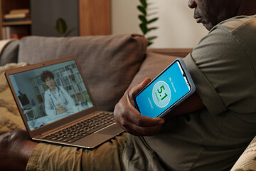 Elderly man having online medical appointment with doctor visible on laptop screen while managing...