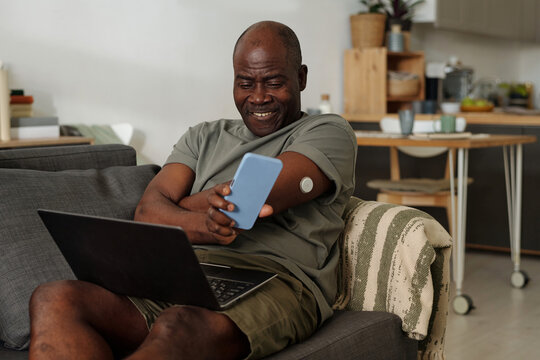 Elderly African American man smiling while checking his smartphone, sitting comfortably on couch with laptop nearby, living room background, insulin monitor on arm