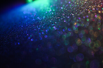 An out of focus, shallow depth of field photograph of a pale light multicolour abstract glittery surface, with vibrant neon light against a black background. Bokeh lights background
