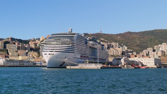 The luxury cruise ship MSC World Europa docked at Genoa&rsquo;s harbor, with the scenic hillside city and clear blue sky forming a dramatic Mediterranean backdrop.