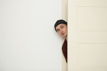 A person with a beanie is curiously peeking around an open door, exhibiting a surprised expression....