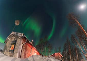 Vibrant green northern lights swirling above a snow covered wooden house and trees in the serene landscape of Lappland, Sweden, creating a magical winter spectacle under the moonlight