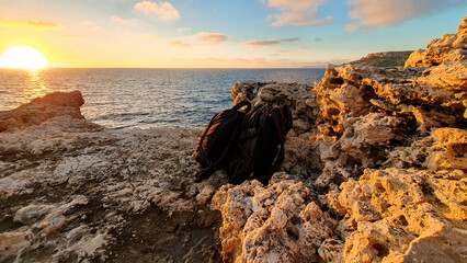 At sunset on the rocky coast of Malta, tourists put down their bags and watch the spectacular...