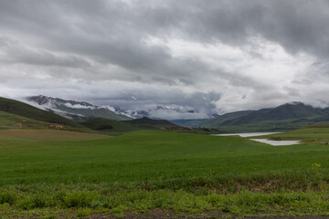 Fototapeta premium A dramatic cloudy landscape near Sisian, Armenia, showcasing lush green meadows, and distant snow-covered peaks partially covered by mist