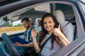 Smiling young woman with license showing thumbs up and driving school instructor with clipboard in car. Driving test, driver courses, exam concept