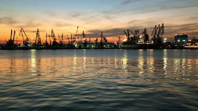 Coucher de soleil observ&eacute; depuis la zone portuaire de Varna, Bulgarie. Les grues du chantier naval voisin se d&eacute;tachent sur le ciel orang&eacute; et se refl&egrave;tent dans les eaux doucement ondulantes de la baie.