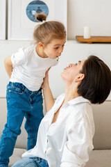 On the couch, a mother and daughter enjoy a tender, playful moment. The woman, dressed in a white shirt with pearls, touches her daughter s nose, evoking a sweet smile.
