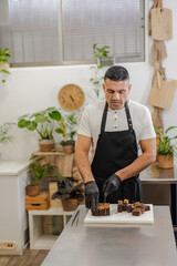 Pastry chef wearing a black apron and gloves carefully slices a chocolate dessert on a white cutting board. Vertical image, blurred background, indoor setting, professional workspace. Copy-space