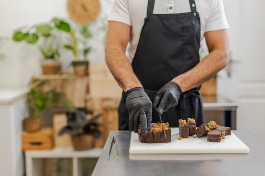Unrecognizable pastry chef, partially visible, slices a chocolate dessert on a white cutting board on the stainless steel table. Horizontal, blurred background, copy-space.