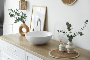 Chest of drawers with sink, photo frame and vases with eucalyptus twigs near white wall in bathroom