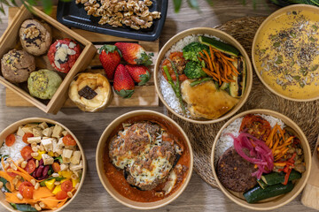 Top-down view of fresh takeaway meals, salads, and desserts in eco-friendly bowls on a wooden table. Includes cookies, cheesecake, salads and fresh fruit. Green plants frame the image, natural touch.