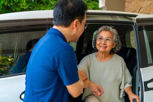 Asian man caregiver or taxi driver helping senior woman patient get in car to get to medical appointments or healthcare therapy sessions at hospital. Assisted elderly patient transportation services.